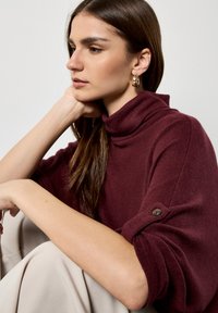 Suéter burdeos de cuello alto con una textura suave, cuello enrollado y detalles de botones en las mangas, combinado con pantalones beige claros.