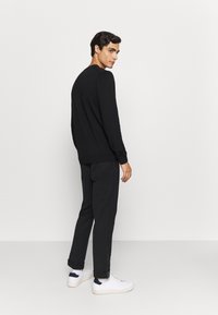 Zwarte lange mouw trui met ribbelboorden, gecombineerd met donkere broeken en witte sneakers met marineblauwe accenten, staand tegen een effen achtergrond.