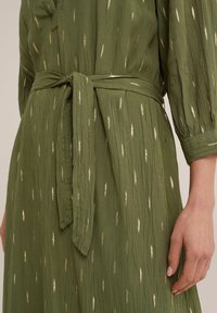 Olivgrön klänning med en texturerad tyg, prydd med metalliska guld vertikala ränder och ett knytbälte i midjan. Långa ärmar med samlade manschetter.