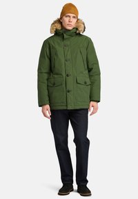Groene geïsoleerde parka met een capuchon van nepbont, sluiting met knopen aan de voorkant en meerdere klepzakken. Gecombineerd met donkere jeans en zwarte schoenen.