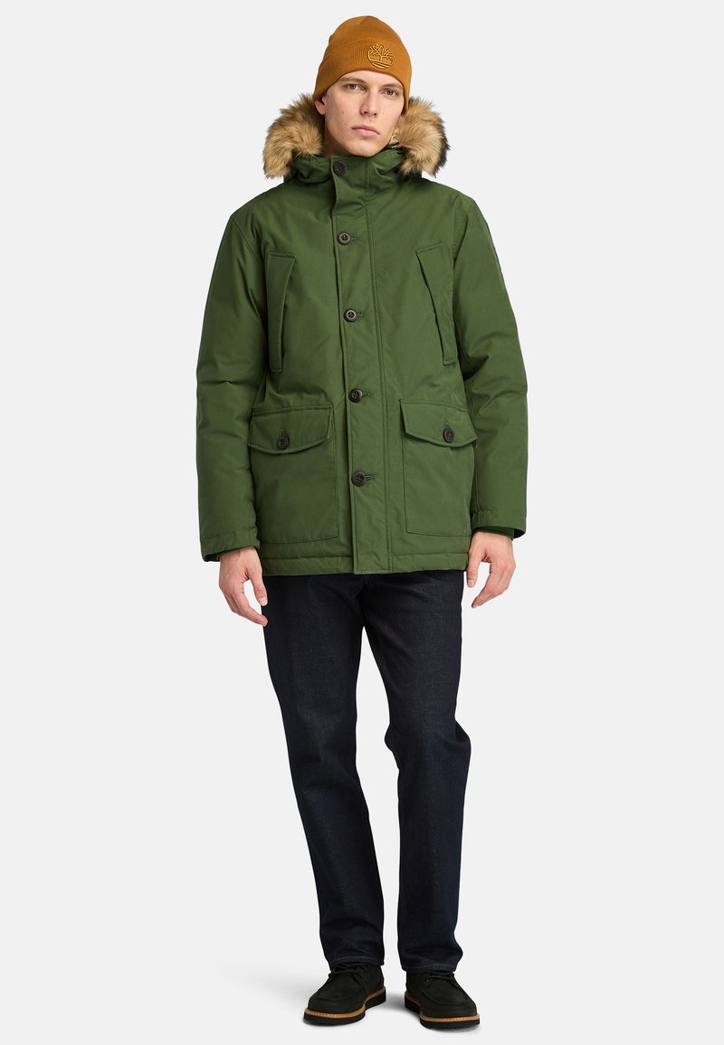 Parka verde isolata con cappuccio foderato in pelliccia sintetica, chiusura con bottoni frontali e molteplici tasche a pattina. Abbinata a jeans scuri e scarpe nere.