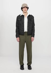 Svart skinnjakke med to frontlommer, hvit skjorte under, olivengrønne bukser og en beige caps. Modellen står mot en hvit bakgrunn.