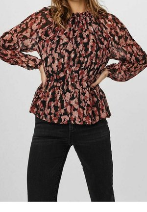 Mujer con blusa de manga larga negra con patrón floral rosa y pantalones negros, con las manos apoyadas en las caderas sobre un fondo liso.