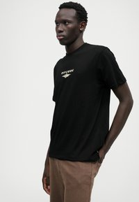 T-shirt de algodão preto com um gráfico de logotipo frontal em cores pastel, decote redondo e mangas curtas. Usado com calças castanhas.