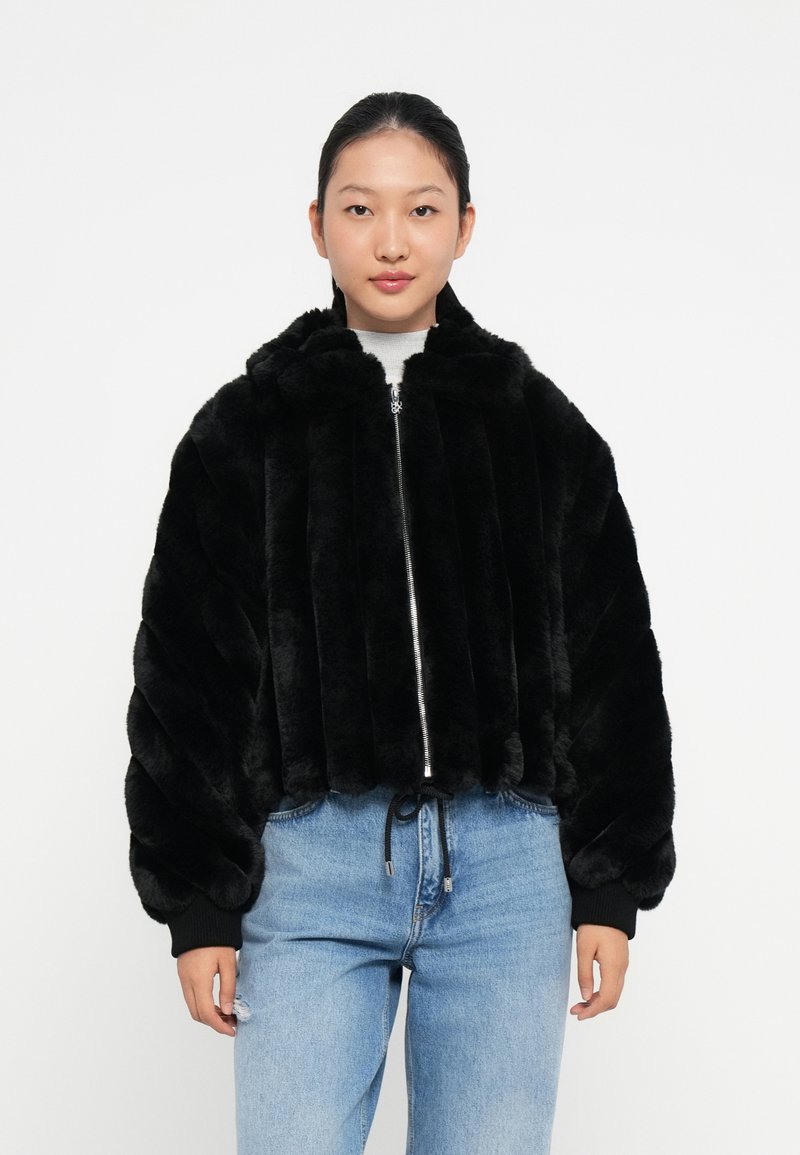 Sort faux fur bomberjakke med lynlås foran, hætte og elastiske manchetter. Designet har vertikale striber og er sat sammen med lyseblå jeans.