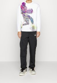 Weißes Langarmshirt mit einem bunten Grafikdesign; kombiniert mit schwarzen Cargo-Hosen und grauen Sneakers mit weißen Sohlen.
