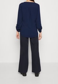 Personne portant un chemisier bleu marine à manches longues et un pantalon large à fines rayures noires, debout de dos sur un fond clair uni.