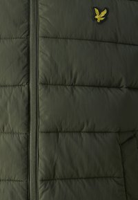 Doudoune vert olive avec un design matelassé, fermeture éclair frontale et un logo d'aigle brodé en haut à gauche.