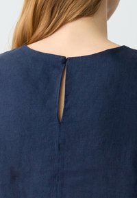 Marineblaues Leinenoberteil mit Rundhalsausschnitt, ausgestattet mit einem kleinen Rückenschlitz, der mit einem Knopf gesichert ist. Sanfte Textur, minimalistisches Design.