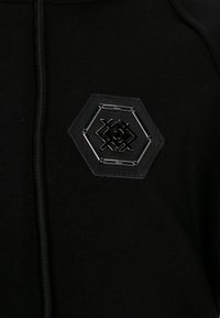 Schwarzer Hoodie mit einem sechseckigen, strukturierten Patch mit glänzendem schwarzen Design auf der Brust. Der Stoff wirkt weich und langlebig.