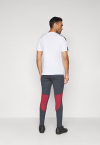 Chemise blanche à manches courtes avec des accents noirs, associée à un legging gris avec des panneaux rouges. Positionnée de l'arrière, mettant en valeur la tenue.