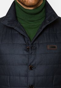 Kurtka w ciemnogranatowym kolorze, pikowana, z zapinaniem na guziki, z teksturą w jodełkę i małą brązową metką marki po lewej stronie klatki piersiowej.