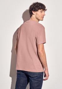 Camiseta de manga corta, cuello redondo, color rosa claro, hecha de una tela suave, llevada con pantalones vaqueros de mezclilla azul oscuro, observada desde atrás.