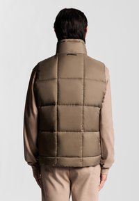 Puffweste in gedämpftem Braun, mit gesteppten, gequilteten Einsätzen. Hoher Kragen und glatte, glänzende Stoffoberfläche. Minimalistisches Design, keine sichtbaren Beschläge.