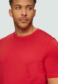 Man met kort krullend haar en een baard, gekleed in een effen rode s.Oliver ronde hals t-shirt, die zijwaarts kijkt tegen een lichte achtergrond.