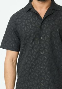 Chemise noire à manches courtes avec un motif texturé de formes géométriques, dotée d'une fermeture à boutons sur le devant et d'un col.