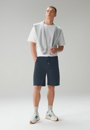 Junger Mann steht mit den Händen in den Taschen, trägt ein weißes T-Shirt, marineblaue Shorts, weiße Turnschuhe, Socken und einen grauen Pullover, der über die Schultern gelegt ist.