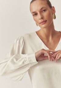 Kremasta bluza z globokim V-izrezom, napihnjenimi dolgimi rokavi in zapenjanjem na gumbe na rokavih. Gladek tekstura, preprost dizajn in nevtralna barva.