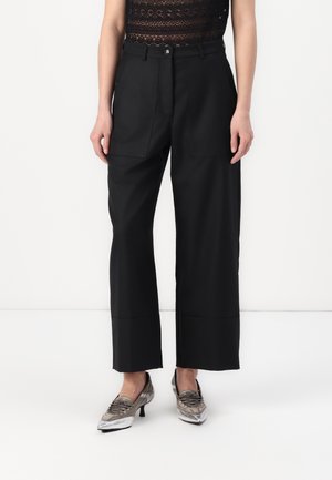 Pantaloni neri a gamba larga con tasche frontali, vestibilità sartoriale, texture liscia, abbinati a scarpe argentate metalliche e a un top di pizzo scuro.