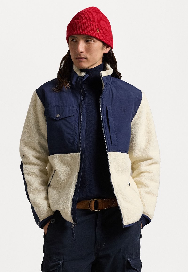 Fleecejakke med marineblå øverste del og cremefarvet nederste del, lynlås lukning, to brystlommer, båret over en marineblå sweater, parret med en rød hue.