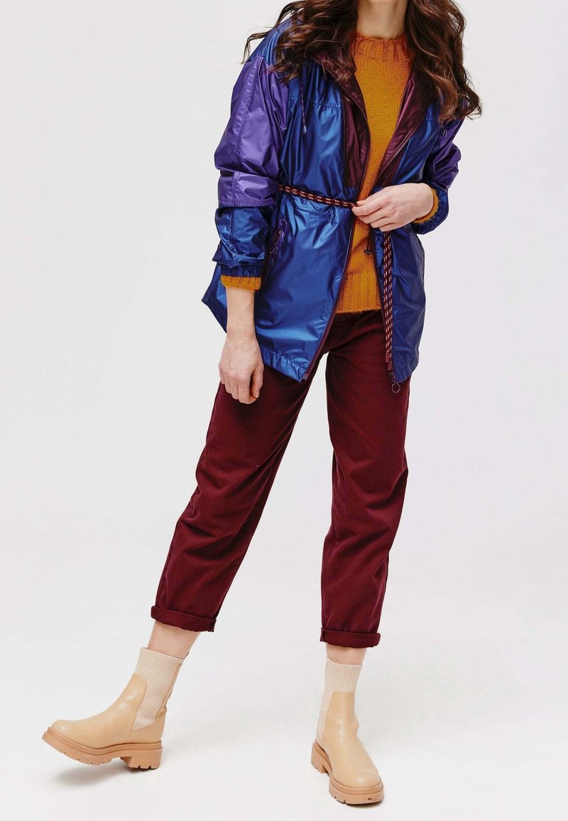 Femme portant une veste bleue brillante, un pull orange, un pantalon bordeaux retroussé et des bottines beiges, debout sur un fond blanc.
