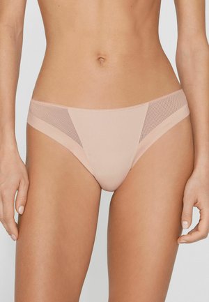 Frau trägt hellbeige, tiefsitzende Bikini-Slips mit Netzeinsätzen an den Hüften vor grauem Hintergrund.