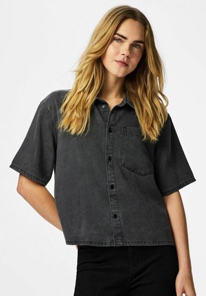 Noor naine, kellel on pikad blondid juuksed, kannab lahtist lühikeste varrukatega musta teksariidest nööpidega särk, seistes ühevärvilise tausta ees.