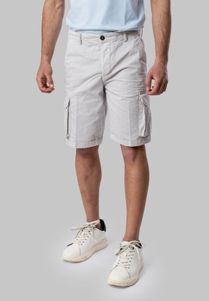 Uomo che indossa pantaloncini cargo beige chiaro e sneakers bianche, in piedi davanti a uno sfondo grigio uniforme.