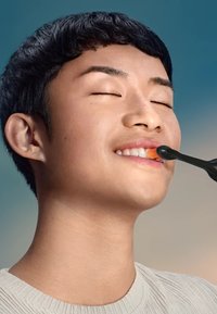 Une brosse à dents électrique avec un manche noir et une tête orange, utilisée par une personne aux cheveux courts et noirs, souriante avec les yeux fermés.