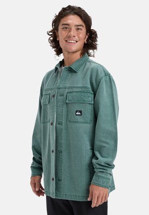 Green denim shirt jacket with a button front, two chest pockets, and a logo patch. Features a classic collar and long sleeves.