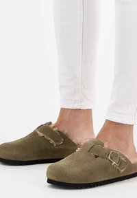 Sabots en suede olive avec une douce doublure en fourrure et un détail de sangle à boucle ; semelle en caoutchouc noire pour l'adhérence ; portés avec un jean skinny blanc.