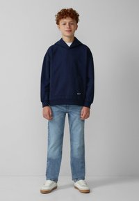 Felpa con cappuccio di colore blu navale, dotata di tasca frontale e polsini a costine, abbinata a jeans dritti di colore blu chiaro e sneakers bianche con dettagli color nocciola.