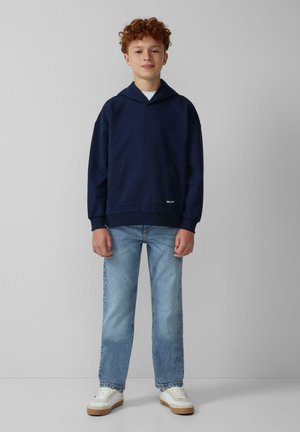 Felpa con cappuccio di colore blu navale, dotata di tasca frontale e polsini a costine, abbinata a jeans dritti di colore blu chiaro e sneakers bianche con dettagli color nocciola.