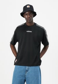 Fekete sportpóló fehér "adidas" logóval, három fehér csíkkal a vállakon. Világoskék farmer rövidnadrággal párosítva.