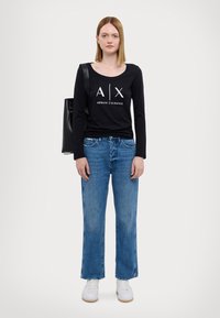 Zwarte langemouwen shirt met witte tekst "A|X" en "ARMANI EXCHANGE", gecombineerd met blauwe wijde jeans en witte sneakers. Zwarte handtas over de schouder gehangen.