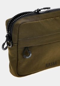Sacca in nylon verde oliva con zip nera e tiranti. Presenta dettagli di cucitura sottili e un logo impresso sul lato. Compatta e rettangolare.