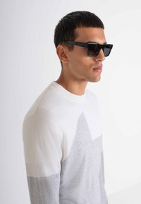 Maglione a maglia bianco con motivo geometrico grigio; presenta un collo rotondo a coste. Il modello indossa occhiali da sole neri rettangolari.
