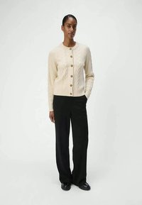 Cardigan en tricot couleur crème avec un motif texturé, comportant une rangée de boutons ronds marron et des bords côtelés, associé à un pantalon noir.