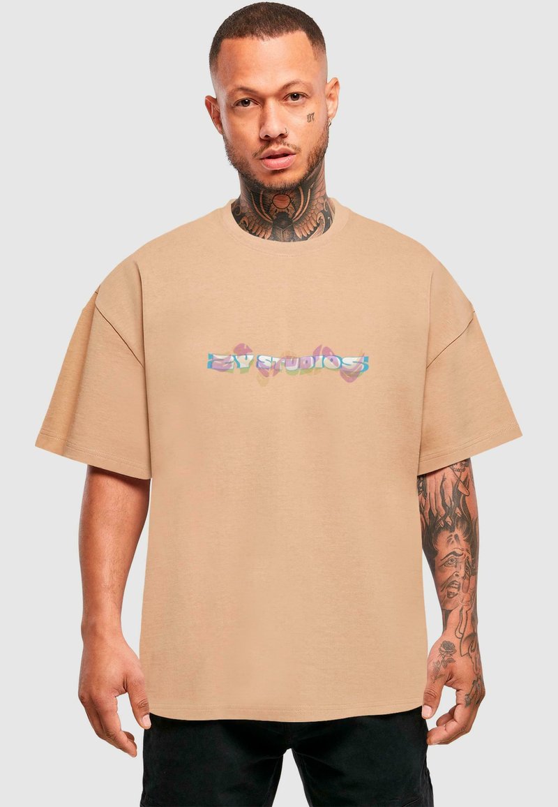 Beige oversized tričko vyrobené z měkkého materiálu, s vícolor grafickým logem "EZY STUDIOS" přes hruď. Krátké rukávy, kulatý výstřih.