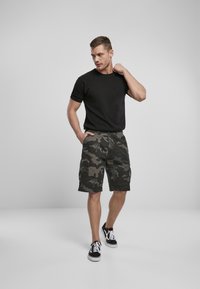 Zwarte t-shirt gecombineerd met camo cargoshorts in donkergroen en grijs, met meerdere zakken. Model draagt zwarte sneakers.