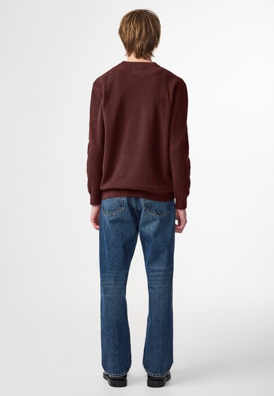 Burgunderfarbener Strickpullover mit geripptem Muster, Rundhals und langen Ärmeln. Kombiniert mit blauen Jeans, die am Saum leicht ausgestellt sind.