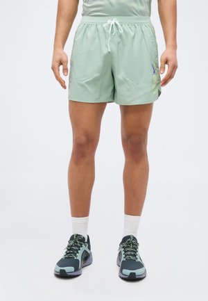 Mees, kes kannab helerohelisi sportluspükse valge paelaga, valgeid sokke ja musta-rohelisi jooksujalatseid, seisab valgel taustal.