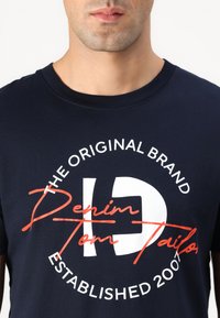 Marineblå bomull t-skjorte med rund hals som har hvit og oransje tekst: "DEN ORIGINALMERKET DENIM TOM TAILOR ETABLERT 2007."