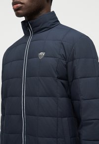 Άνδρας με σκούρο μπλε μπουφάν με επένδυση και όρθιο γιακά, με κάθετο φερμουάρ με λευκή λεπτομέρεια και μικρό λογότυπο EA7 στο στήθος.