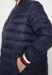 Námořnicky modrá péřová bunda s prošívaným vzorem, zapínáním na zip, žebrovaným límcem a proužkovanými manžetami. Obsahuje boční kapsy a nenápadný detail značky.