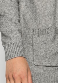 Šedý pletený svetr s vroubkovanými manžetami a přední kapsou, který vyniká měkkou texturou a uvolněným střihem.