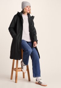 Woman wearing a gray knit hat, black long coat, light sweater, blue jeans, and white sneakers, sitting on a wooden stool against a plain background.