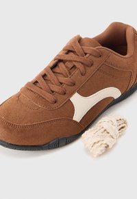 Καφέ σουέτ αθλητικό παπούτσι με λευκή καμπυλωτή λεπτομέρεια, στρογγυλεμένη μύτη, μαύρη λαστιχένια εξωτερική σόλα και κορδόνια στο ίδιο καφέ χρώμα. Περιλαμβάνει και κρεμ κορδόνι.