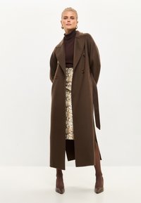 Long manteau marron avec un design croisé, fente sur le côté et ceinture à nouer. Porté sur un col roulé marron et une jupe à motif. Talons marron.