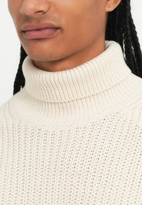 Maglione a collo alto color crema con un motivo di maglieria testurizzato, caratterizzato da un colletto a coste e un tessuto morbido e spesso.